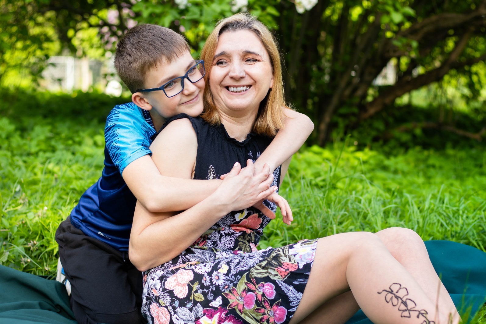 Uśmiechnięta kobieta w kwiecistej sukience siedzi na trawie, obejmowana przez chłopca w okularach i niebieskiej koszulce; w tle zieleń drzew i białe kwiaty.
