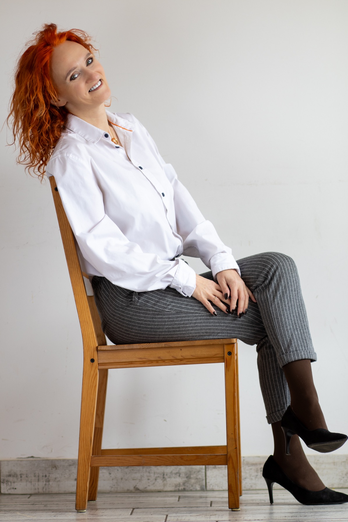 Uśmiechnięta kobieta z rudymi kręconymi włosami, ubrana w białą koszulę i szare spodnie w prążki, siedzi na drewnianym krześle na tle białej ściany.