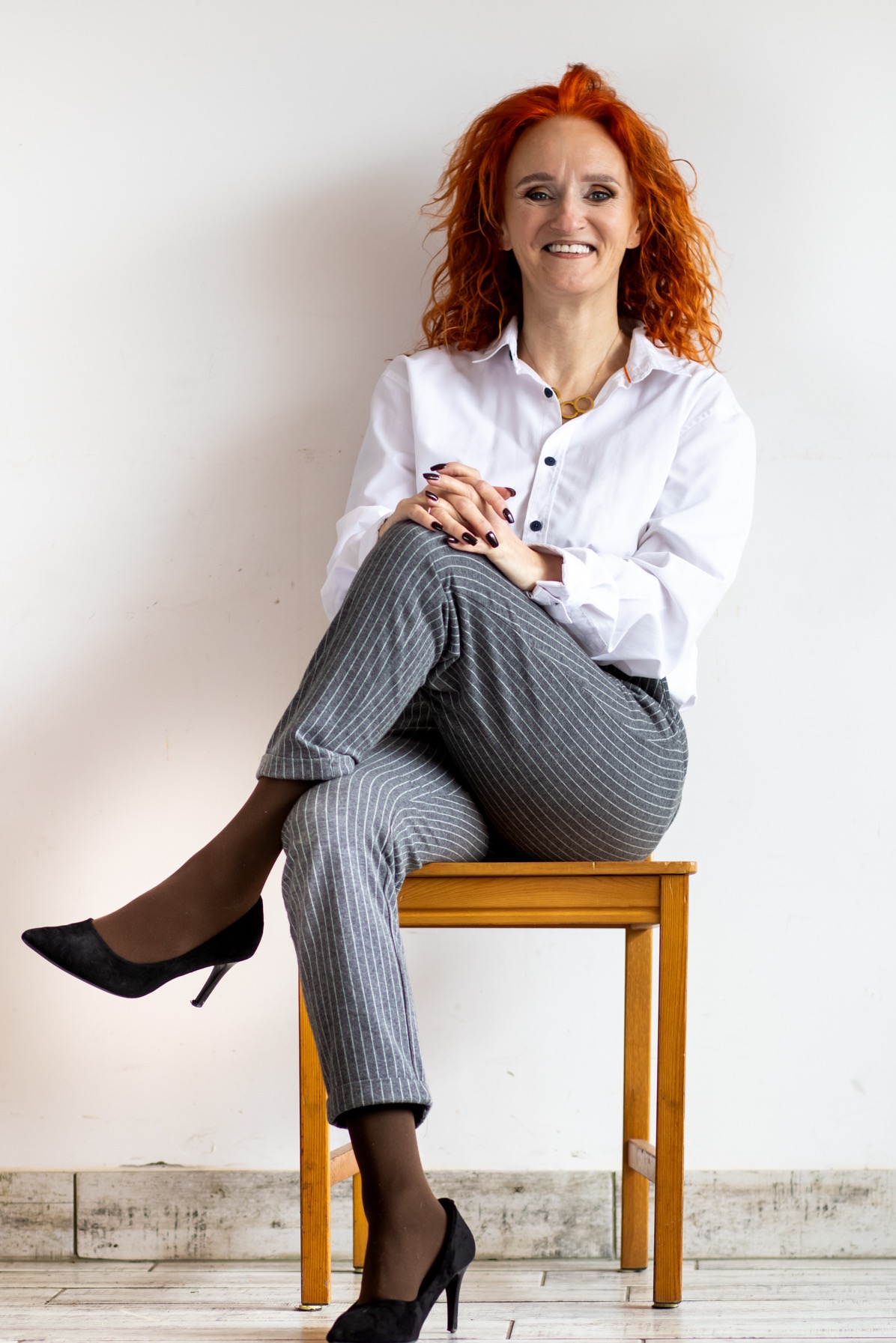 Kobieta z rudymi włosami w białej koszuli i szarych spodniach w paski, siedząca na drewnianym krześle na tle białej ściany, z rękami splecionymi na kolanach i czarnych szpilkach.