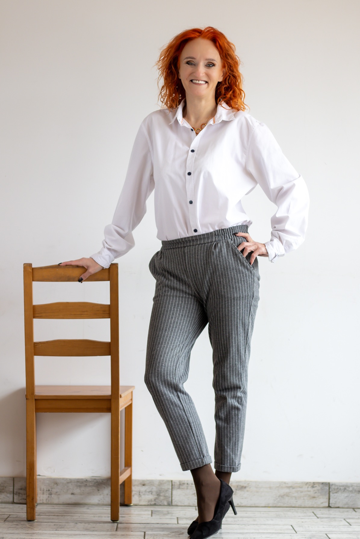 Kobieta z rudymi kręconymi włosami, w białej koszuli i szarych spodniach w paski, pozuje opierając się o drewniane krzesło na tle białej ściany.