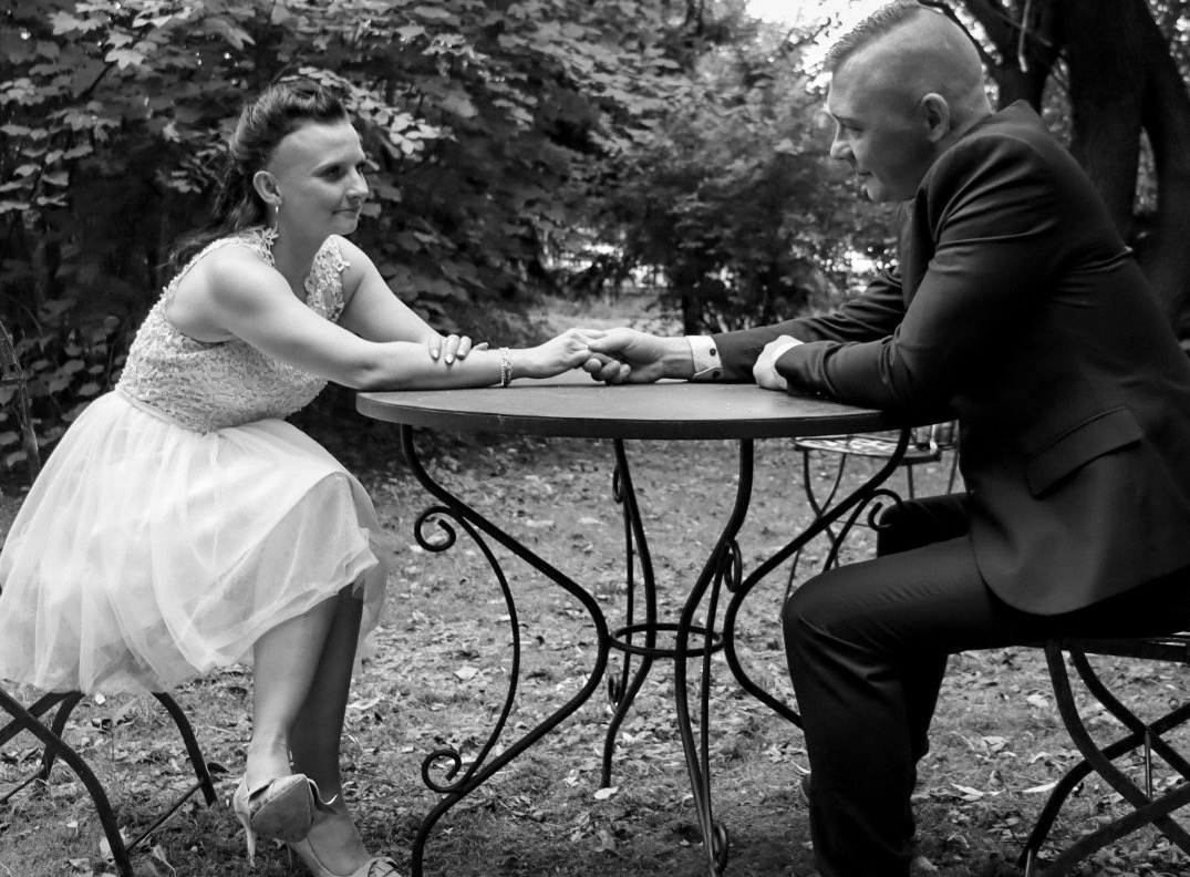 Czarno-białe zdjęcie pary siedzącej przy stoliku w ogrodzie, trzymającej się za ręce: kobieta w sukience z tiulowym dołem i zdobioną górą, mężczyzna w garniturze, oboje zamyśleni.