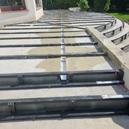 Paweł Lipiński - Konstrukcja tarasu z metalowych legarów, częściowo wypełniona betonem, widoczna drabina i fragment budynku w tle.