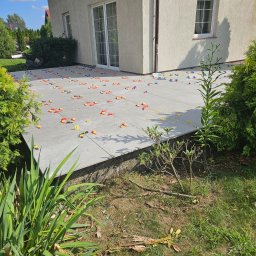 Paweł Lipiński - Ułożony taras z szarych płyt z widocznymi plastikowymi elementami poziomującymi w kolorach czerwonym, żółtym i niebieskim, otoczony zielenią.