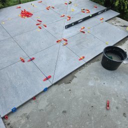 Paweł Lipiński - Układanie szarych płytek na zewnątrz z użyciem poziomicy i klipsów poziomujących w kolorach czerwonym, żółtym i niebieskim, obok stoi czarne wiadro z zaprawą.