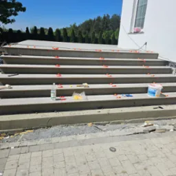 Schody wejściowe do domu w trakcie układania płytek, z użyciem poziomujących klipsów i klinów, widoczne narzędzia i materiały budowlane.