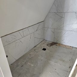 Paweł Lipiński - Fragment łazienki w trakcie remontu: ułożone płytki imitujące marmur na skośnej ścianie poddasza, surowy beton na podłodze z widocznym otworem, fragment futryny drzwi.
