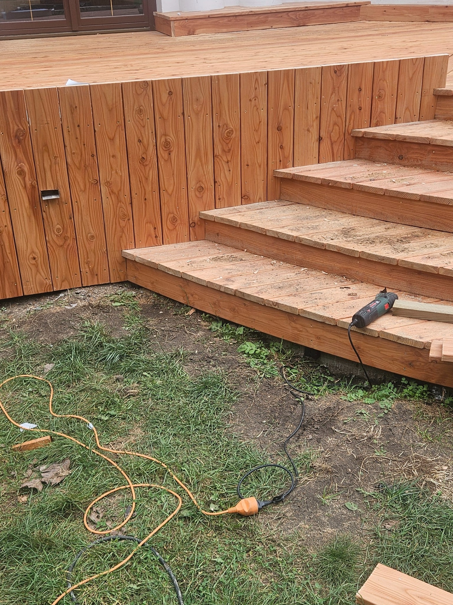Drewniany taras z trzema schodami w trakcie budowy, widoczne narzędzie wielofunkcyjne na schodach i pomarańczowy kabel zasilający na trawie.