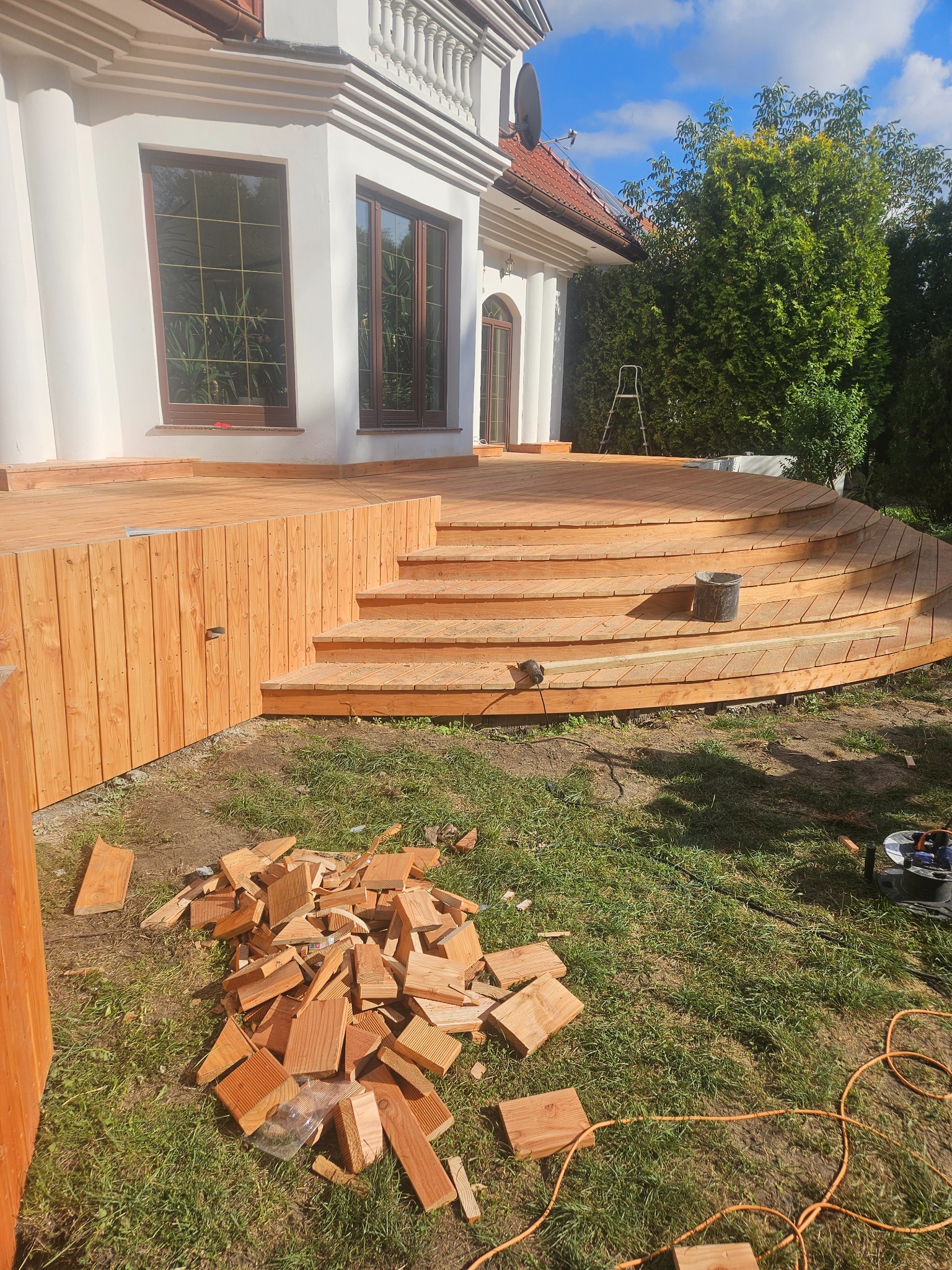 Budowa drewnianego tarasu z zaokrąglonymi schodami przy białym domu, widoczne narzędzia i resztki drewna na trawie.