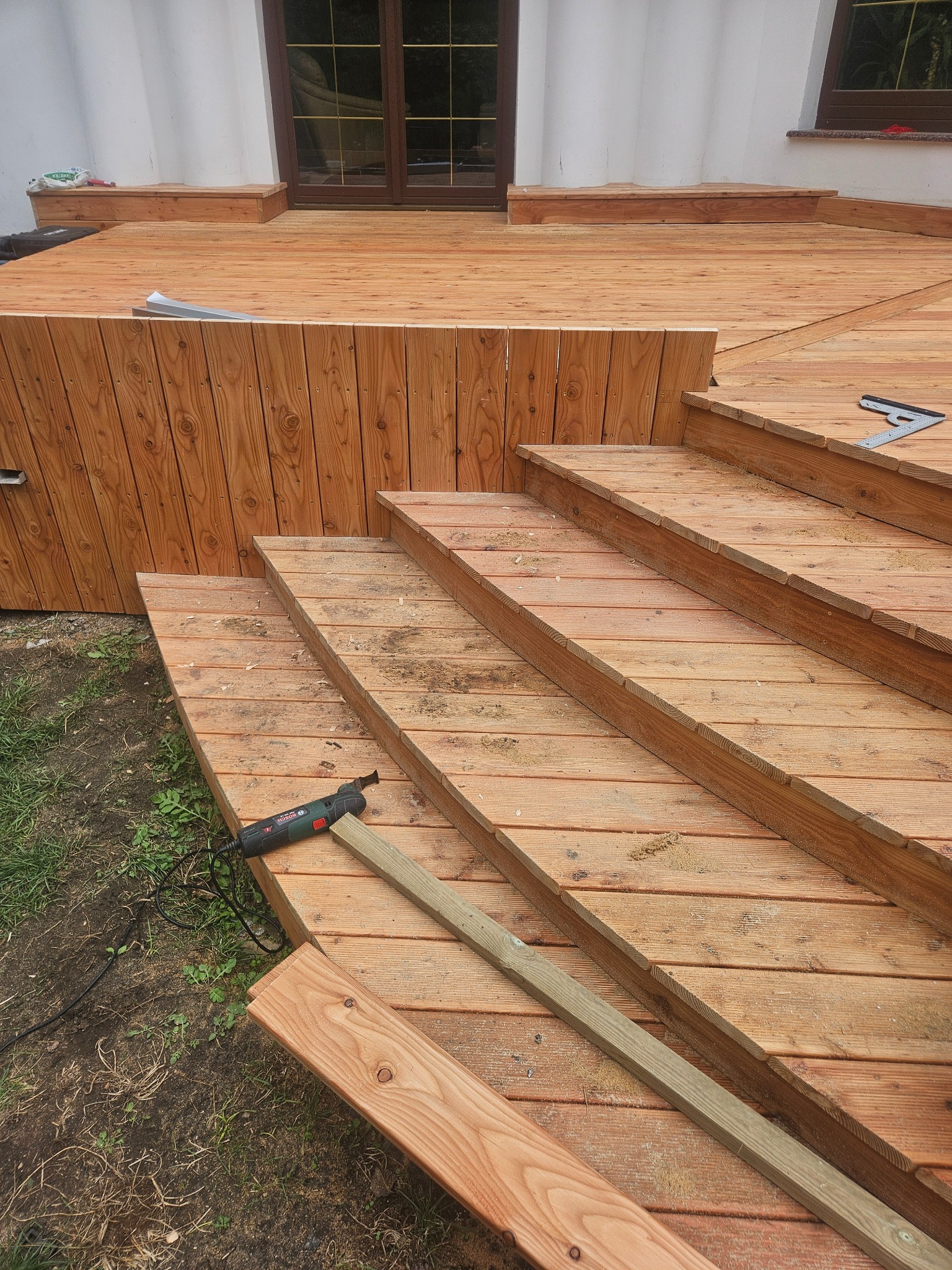 Budowa drewnianego tarasu z schodami, widoczny fragment domu z oknem, narzędzie wielofunkcyjne Bosch, deski i kątownik.