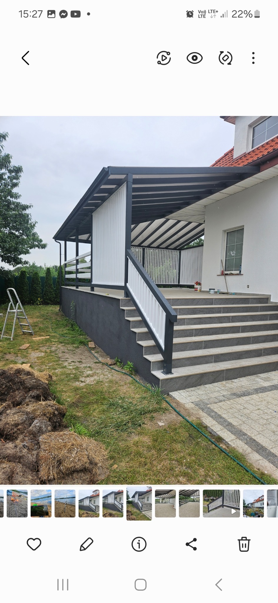 Zadaszony taras z białymi panelami, czarną konstrukcją i schodami z szarego kamienia, widok z zewnątrz na tle trawnika i domu z czerwoną dachówką.