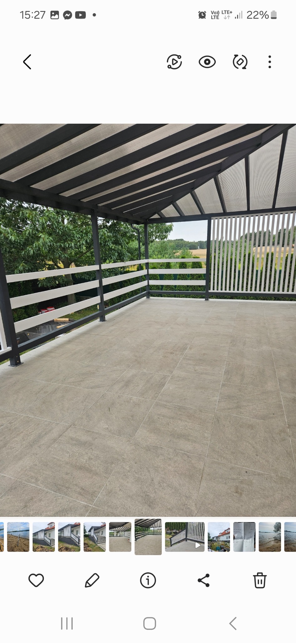 Zadaszony taras z szarą podłogą z płytek, białą balustradą i widokiem na zieleń za nią.