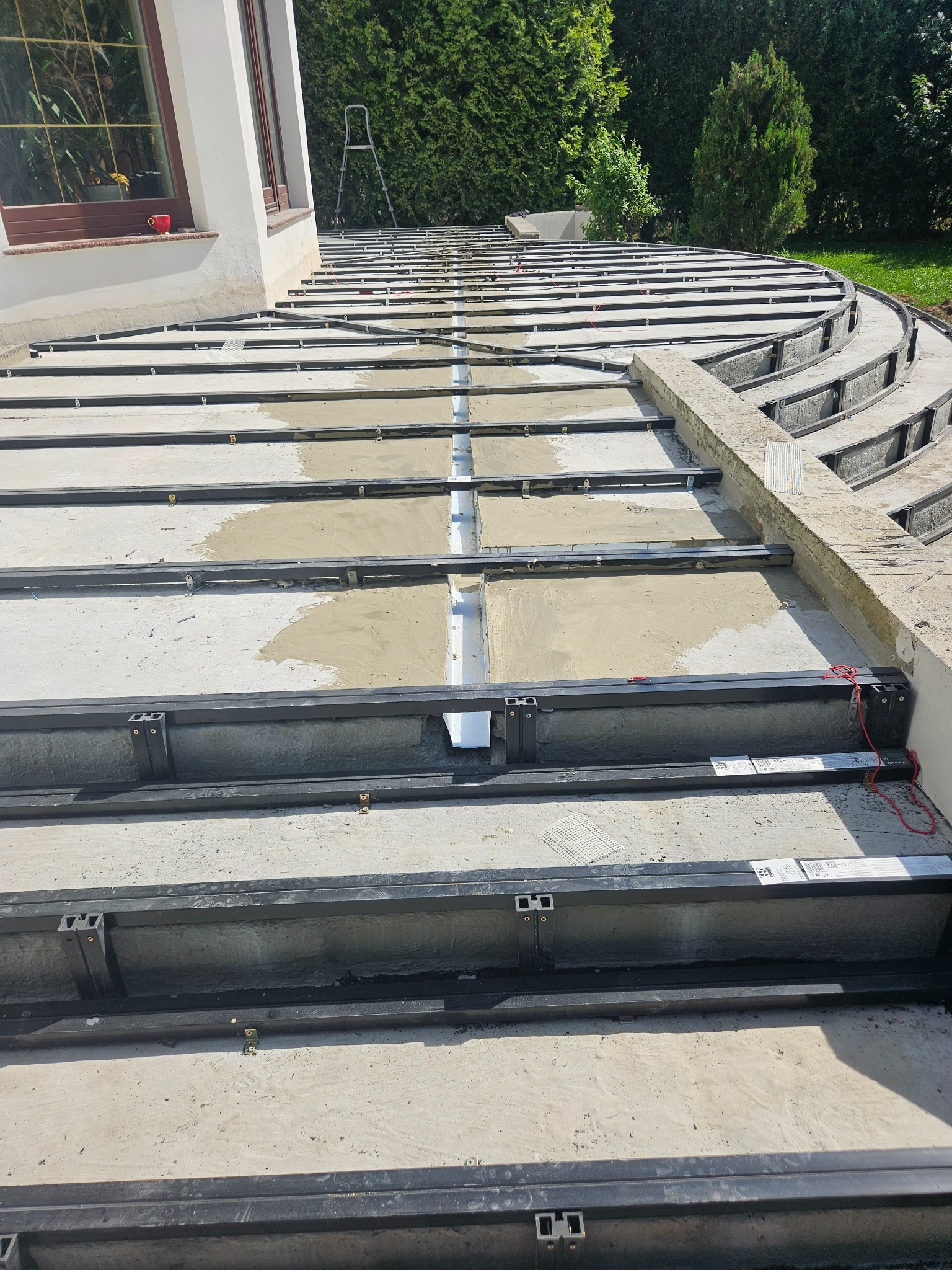 Konstrukcja tarasu z metalowych legarów, częściowo wypełniona betonem, widoczna drabina i fragment budynku w tle.