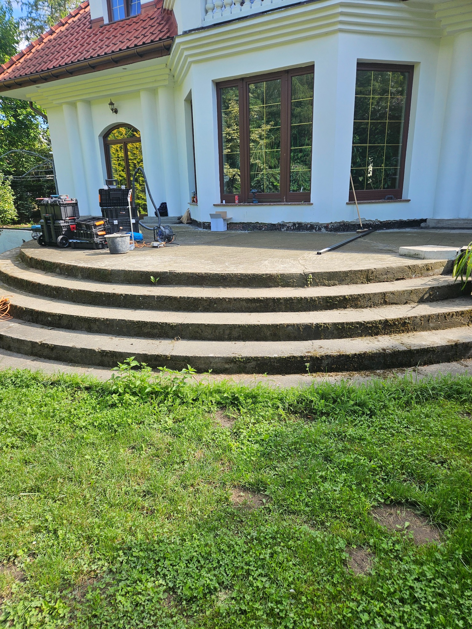 Schody wejściowe do domu z widocznymi śladami prac remontowych, narzędzia i skrzynki narzędziowe obok, trawnik w pierwszym planie.