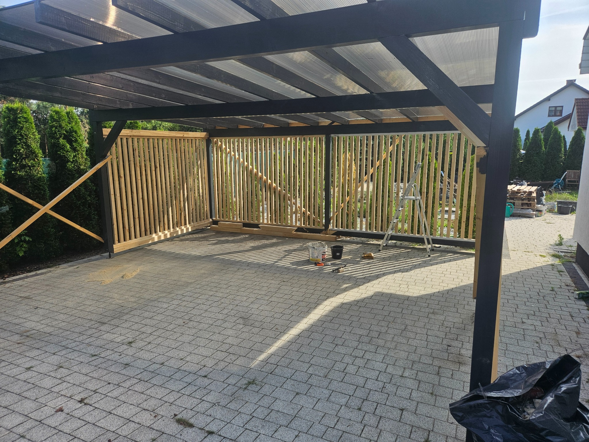 Drewniana konstrukcja z zadaszeniem z poliwęglanu w trakcie budowy, widoczne listwy ogrodzeniowe i drabina, narzędzia malarskie na kostce brukowej.
