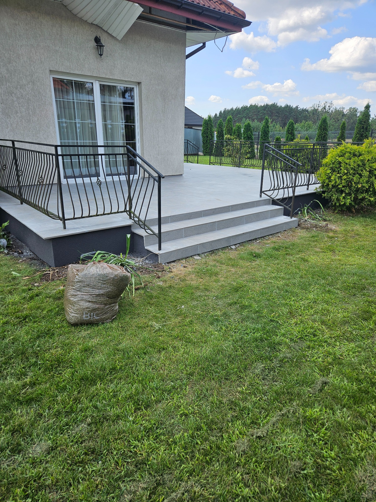 Nowoczesny taras z szarych płytek z czarną balustradą w Toruniu, widok z trawnika na dom z dużymi oknami, obok worka z odpadami ogrodowymi.