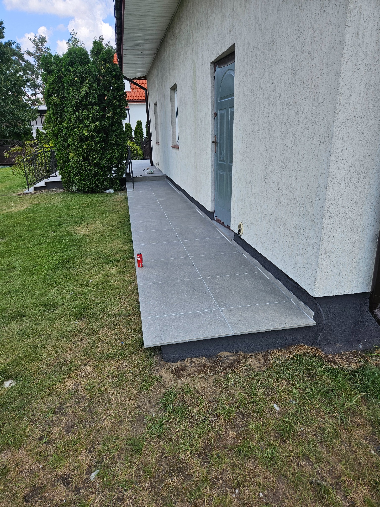 Wykończony taras z szarych płytek przy budynku z widocznym wejściem i puszką napoju na obrzeżu.