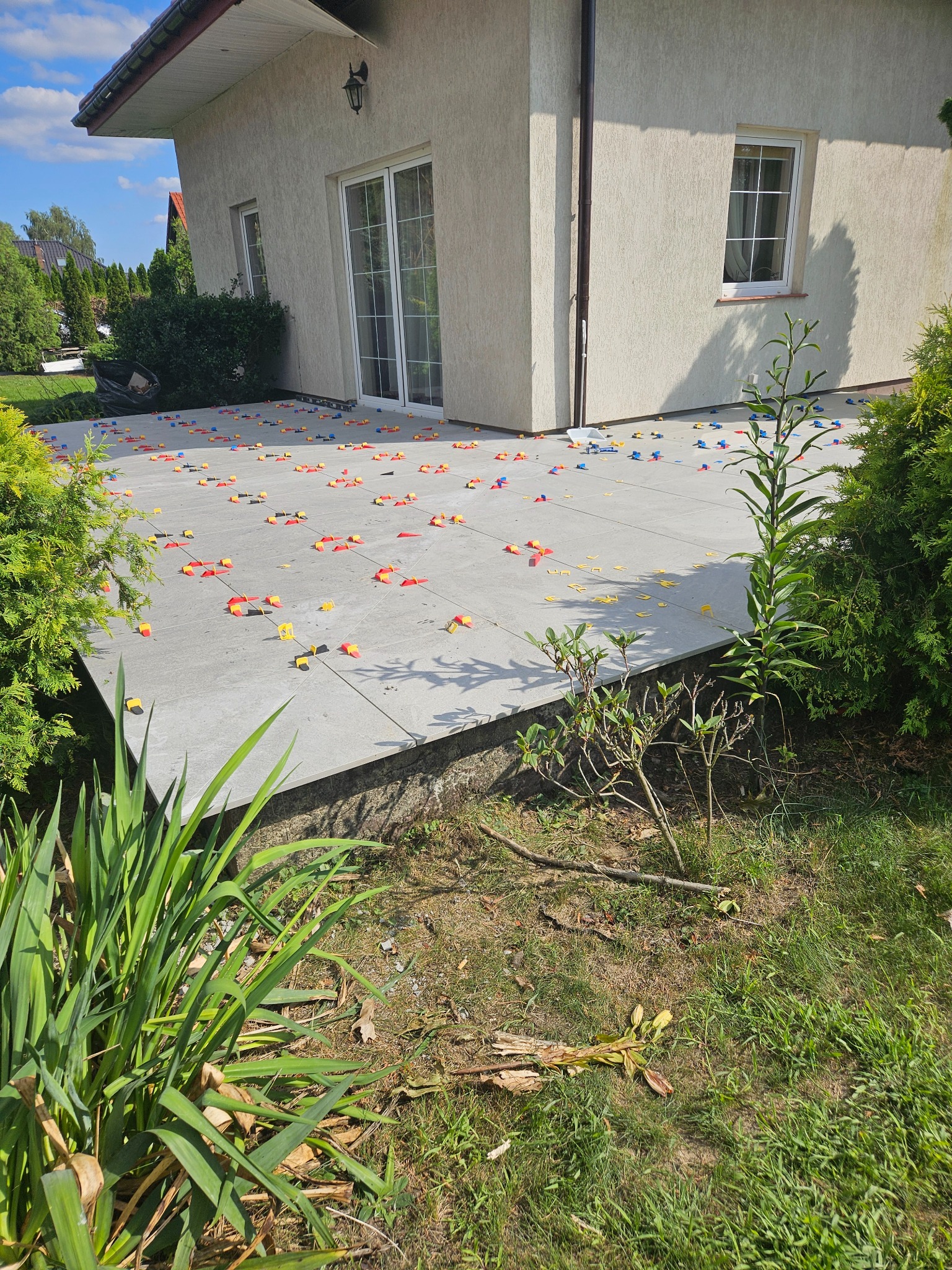 Ułożony taras z szarych płyt z widocznymi plastikowymi elementami poziomującymi w kolorach czerwonym, żółtym i niebieskim, otoczony zielenią.