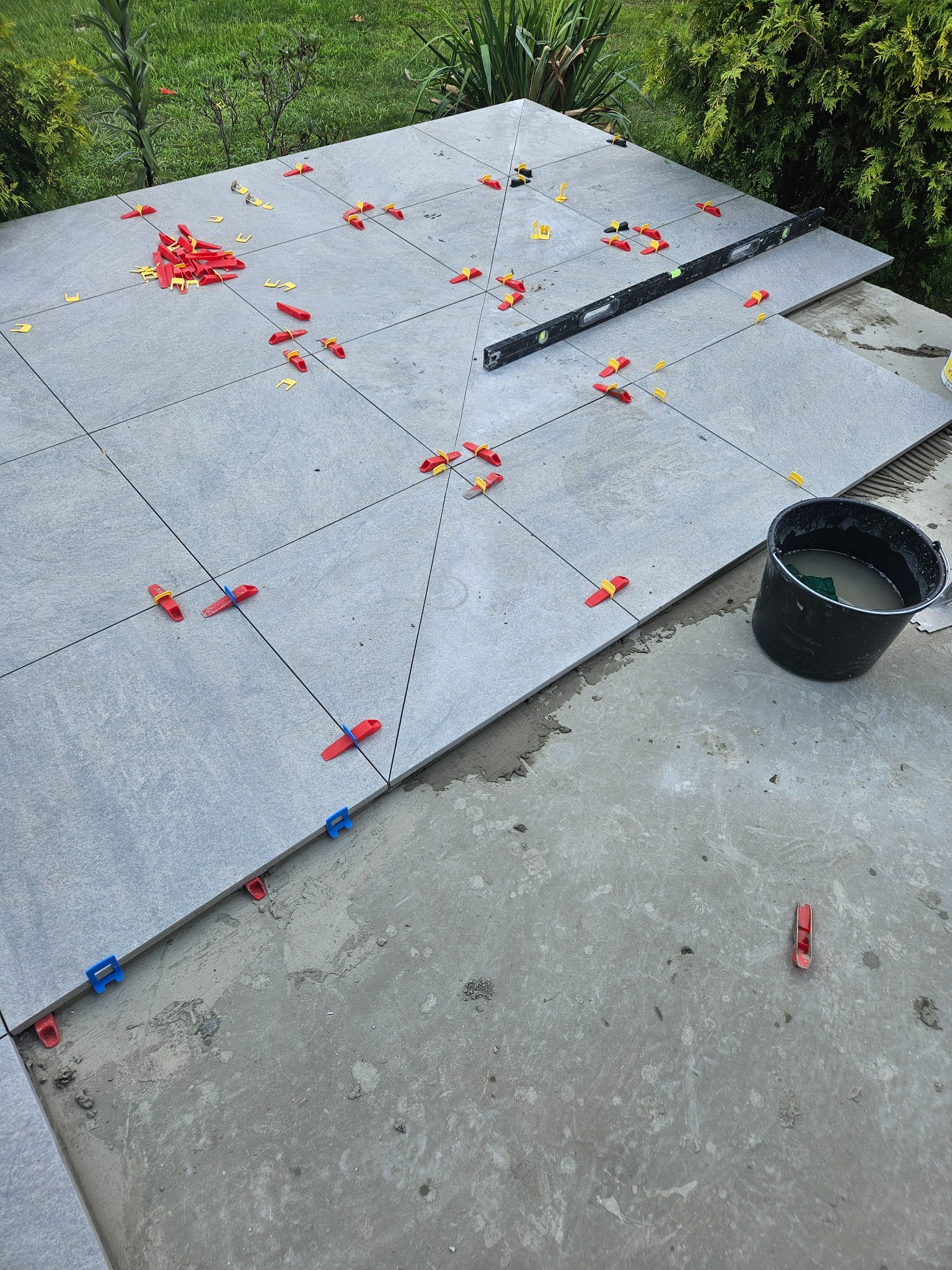 Układanie szarych płytek na zewnątrz z użyciem poziomicy i klipsów poziomujących w kolorach czerwonym, żółtym i niebieskim, obok stoi czarne wiadro z zaprawą.