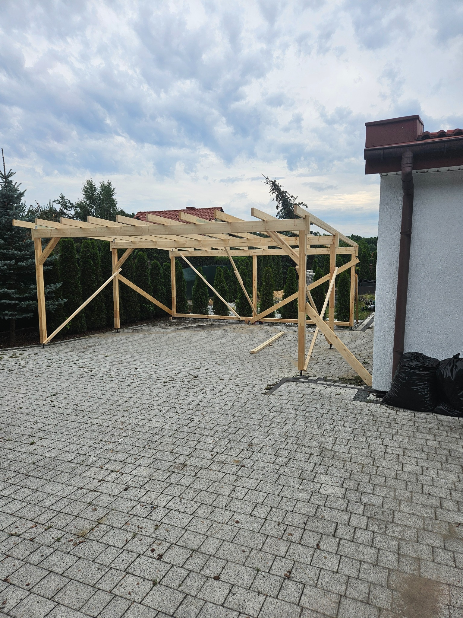 Drewniana konstrukcja z belek, prawdopodobnie zadaszenie tarasu lub wiaty, w trakcie budowy na posesji z kostki brukowej, podparta tymczasowymi ukośnymi belkami, z widocznymi workami na śmieci obok...