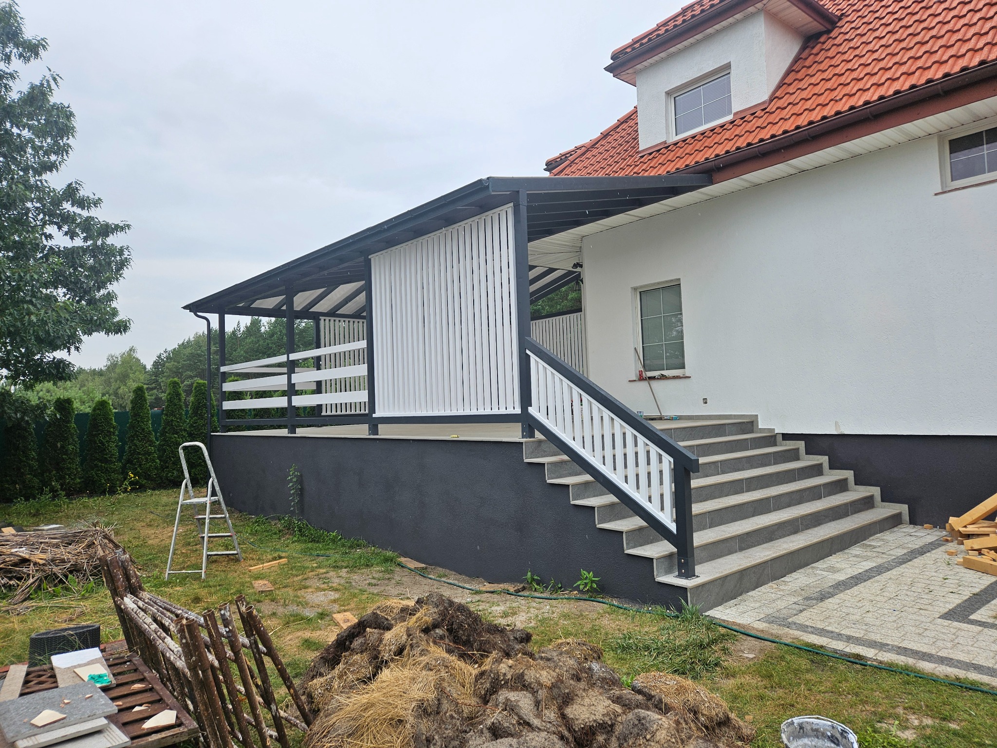 Nowoczesny dom z tarasem z białymi panelami, schodami wyłożonymi szarymi płytkami i czarną podmurówką, widoczny fragment ogrodu z drabiną i materiałami budowlanymi.
