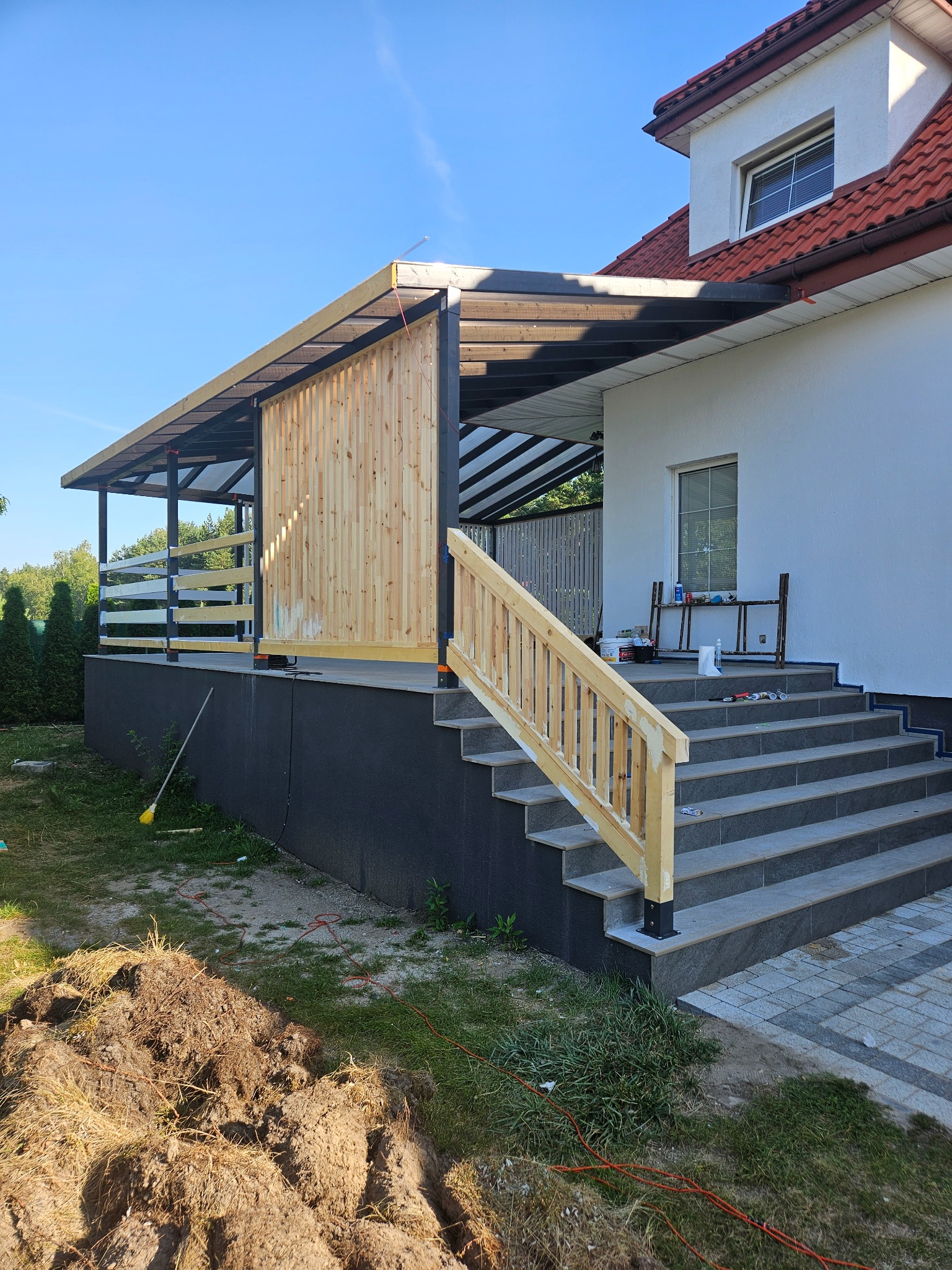 Zadaszony taras z drewnianą ażurową ścianką i nową balustradą w trakcie budowy, widoczne narzędzia i materiały na schodach, świeżo wykopana ziemia obok tarasu.