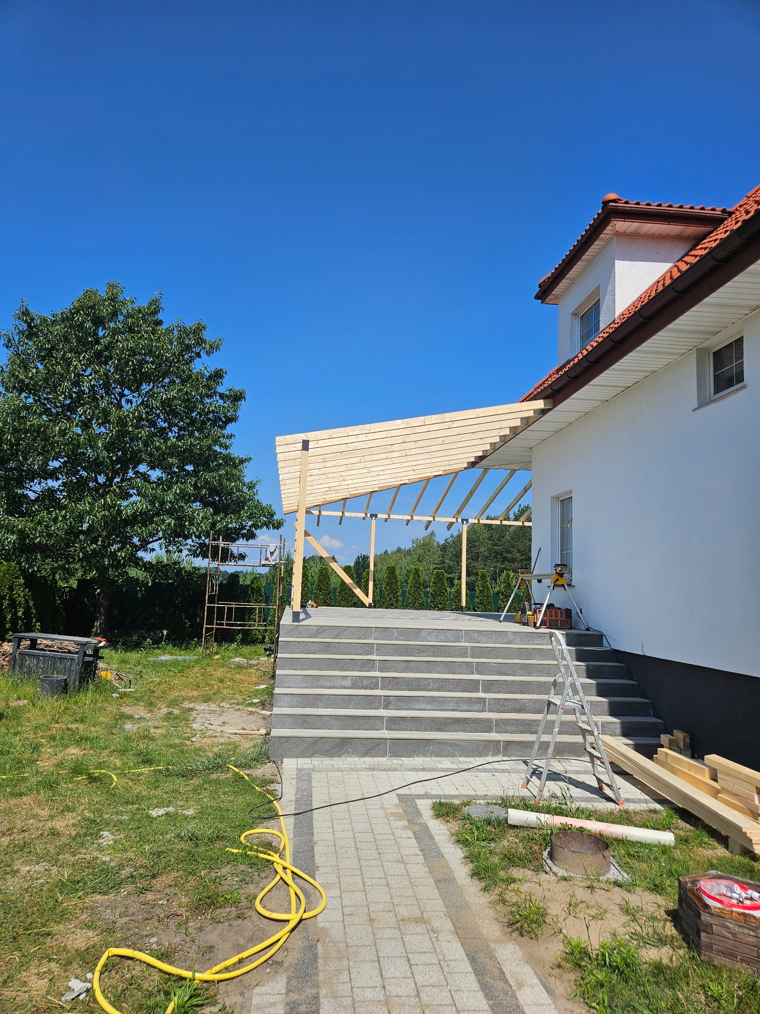 Konstrukcja drewniana zadaszenia tarasu w trakcie budowy, widoczne schody wejściowe do domu, narzędzia i materiały budowlane na zewnątrz.