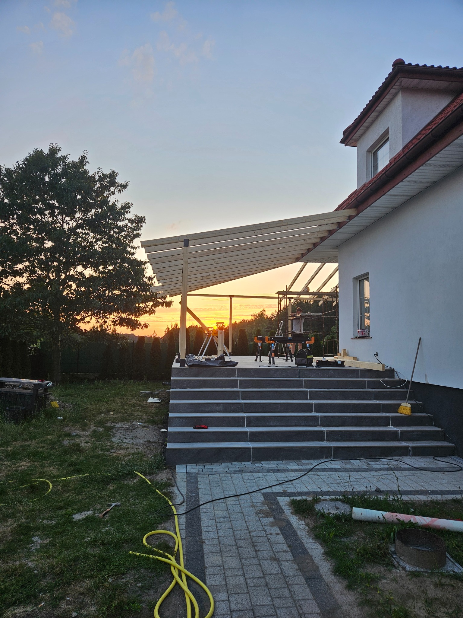 Budowa zadaszenia tarasu przy domu jednorodzinnym, widoczne drewniane elementy konstrukcyjne i narzędzia na tle zachodzącego słońca.