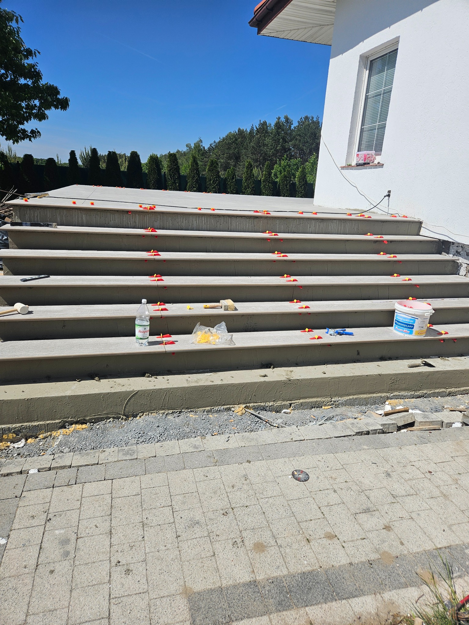 Schody wejściowe do domu w trakcie układania płytek, z użyciem poziomujących klipsów i klinów, widoczne narzędzia i materiały budowlane.