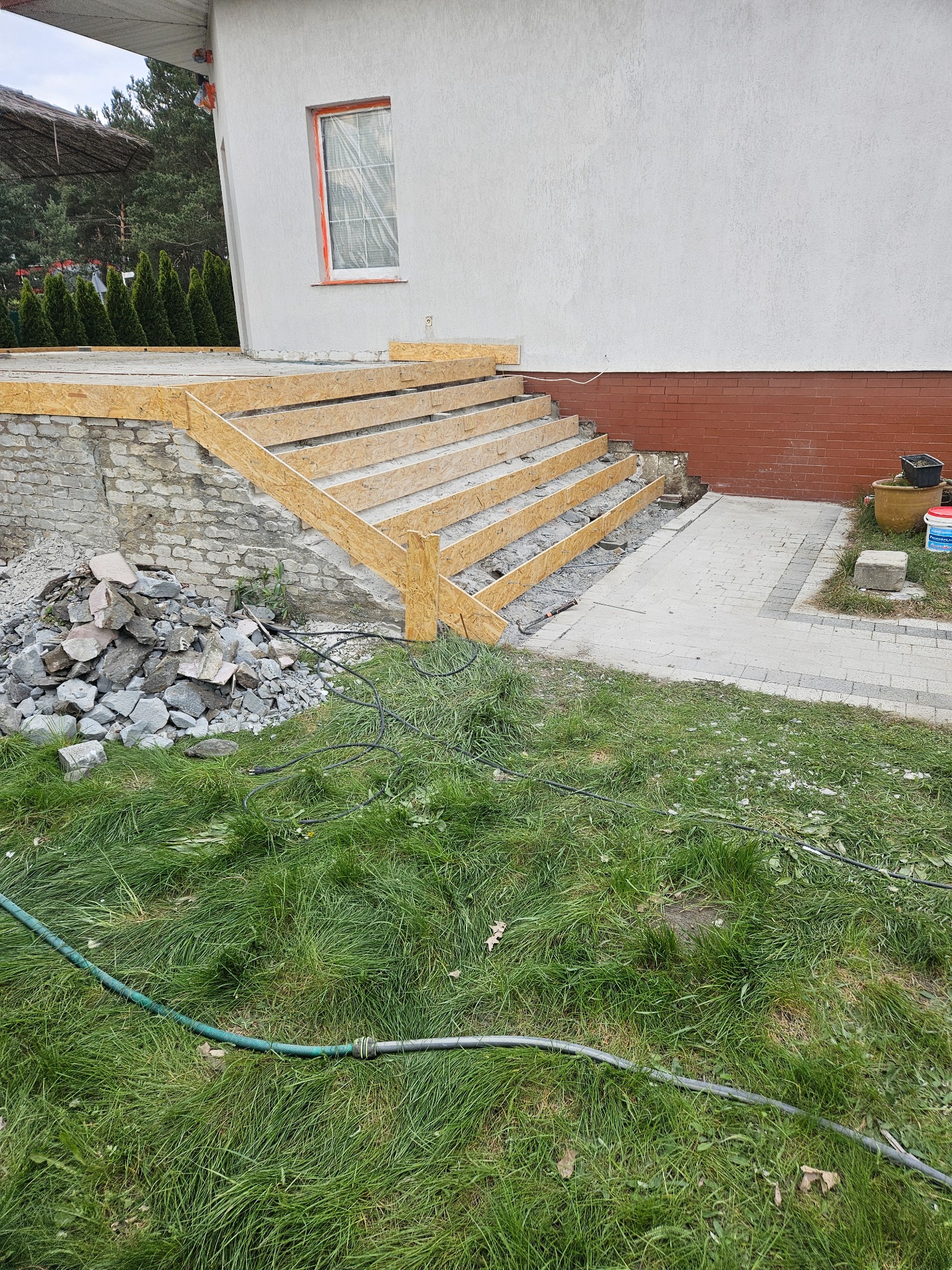 Budowa schodów zewnętrznych z drewnianym szalunkiem przy domu z białą elewacją i ceglanym cokołem, widoczne narzędzia i gruz.