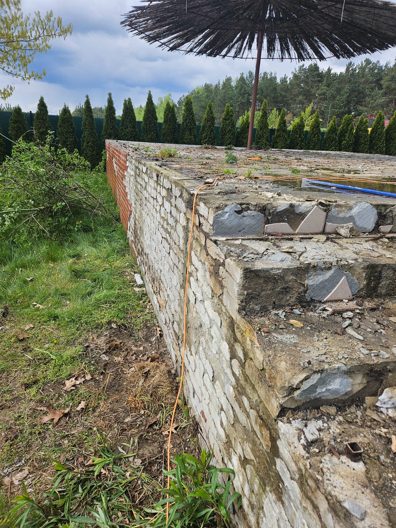 Niedokończona, zniszczona murowana konstrukcja, prawdopodobnie pozostałość po basenie, z widocznymi uszkodzeniami i fragmentami płytek, otoczona trawą i roślinnością, z parasolem trzcinowym w tle.