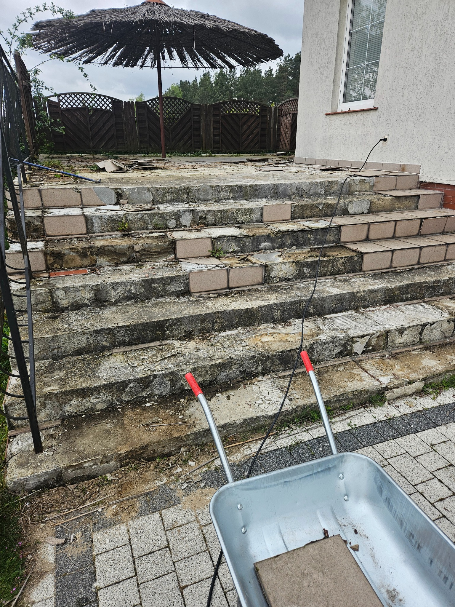 Uszkodzone, betonowe schody zewnętrzne przed budynkiem z widocznymi ubytkami i odspojonymi płytkami, w tle parasol trzcinowy i taczka z fragmentem płytki na kostce brukowej.