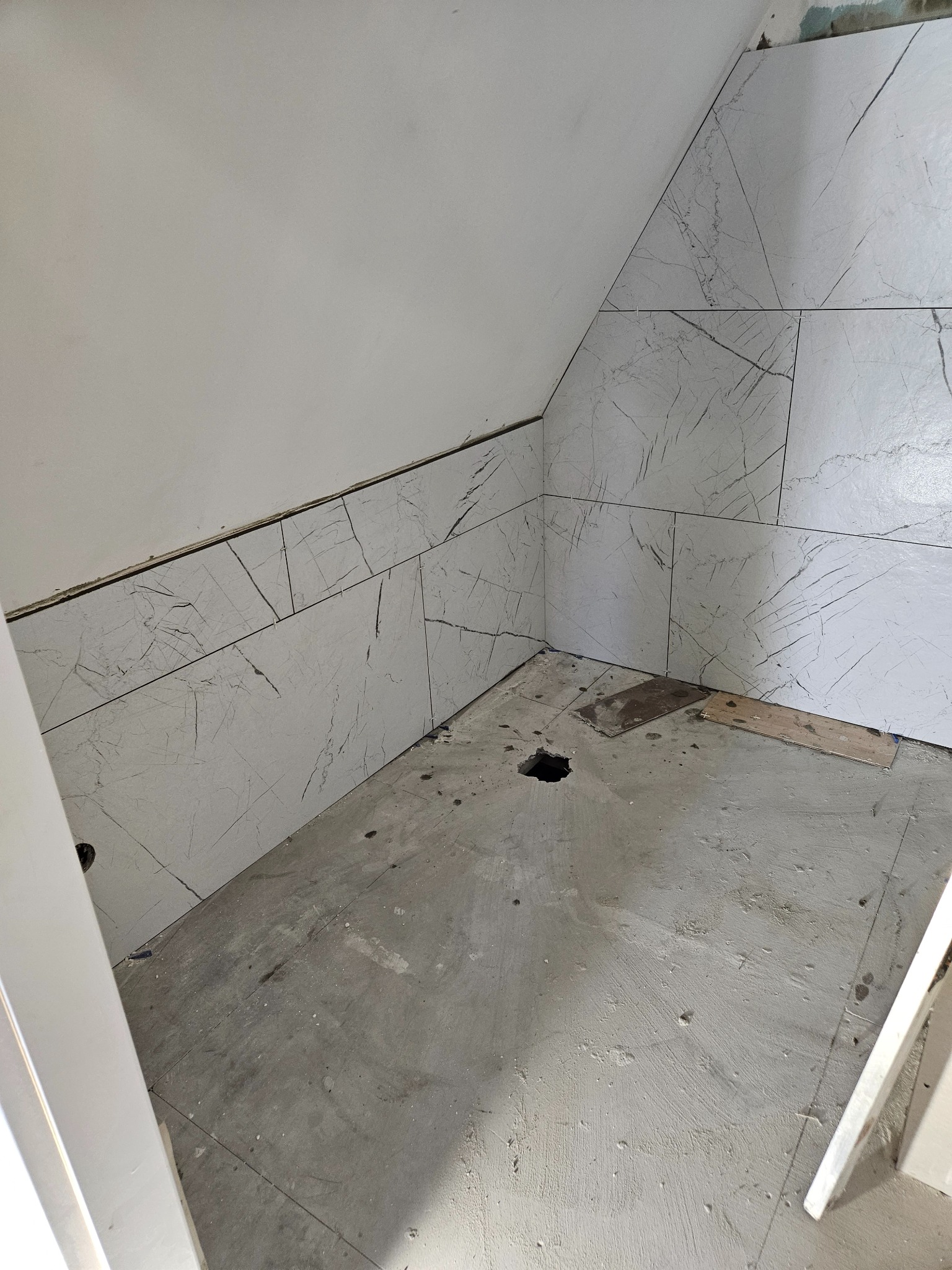Fragment łazienki w trakcie remontu: ułożone płytki imitujące marmur na skośnej ścianie poddasza, surowy beton na podłodze z widocznym otworem, fragment futryny drzwi.