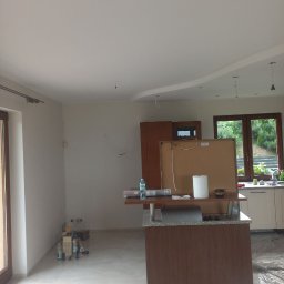 Dmytro Sitalo - Wnętrze kuchni przed wykończeniem: świeżo pomalowane ściany i sufit z dekoracyjnym obniżeniem, widoczne butelki z wodą i materiały budowlane na podłodze, okno z widokiem na zieleń.