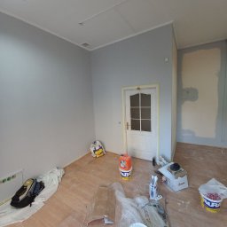 Dmytro Sitalo - Pokój po malowaniu ścian na szaro, z widocznym zabezpieczeniem drzwi taśmą malarską, resztkami farb, narzędziami i folią ochronną na podłodze.