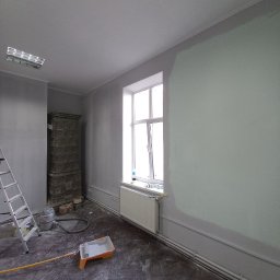 Dmytro Sitalo - Wnętrze pokoju w trakcie odświeżania: ściana częściowo pokryta jasną farbą, widoczny grzejnik, drabina, kuweta malarska z wałkiem, wiadro z farbą i stary piec kaflowy w rogu pomieszczenia.