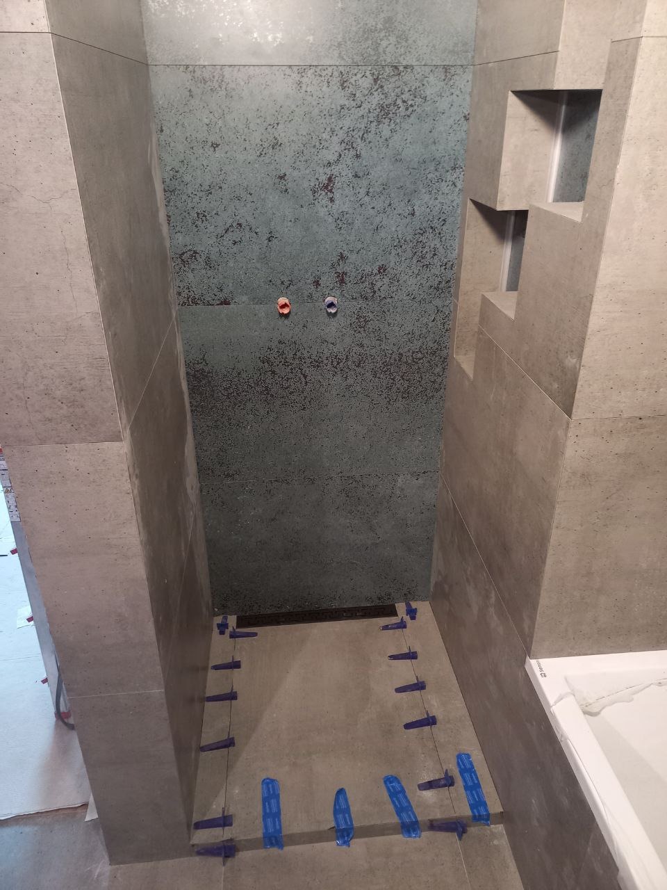 Wnęka prysznicowa w trakcie wykańczania, z szarymi płytkami imitującymi beton, niebieskimi krzyżykami dystansowymi i metaliczną ścianą akcentującą z widocznymi przyłączami wodnymi.