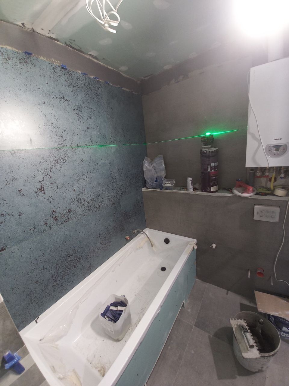 Łazienka w trakcie remontu: wanna obudowana płytami, ściany częściowo pokryte płytkami o wzorze przypominającym beton, widoczny laser krzyżowy, pojemnik z krzyżykami dystansowymi w wannie, wiadro...