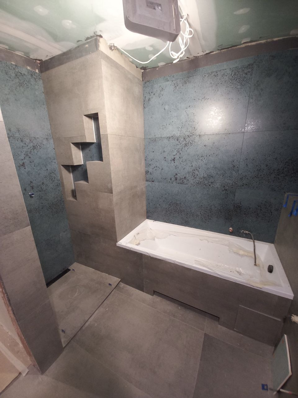 Wnętrze łazienki w trakcie wykańczania: szare płytki na podłodze i ścianach, wanna zabezpieczona folią, widoczne niedokończone prace na suficie, wnęka w ścianie wyłożona płytkami.