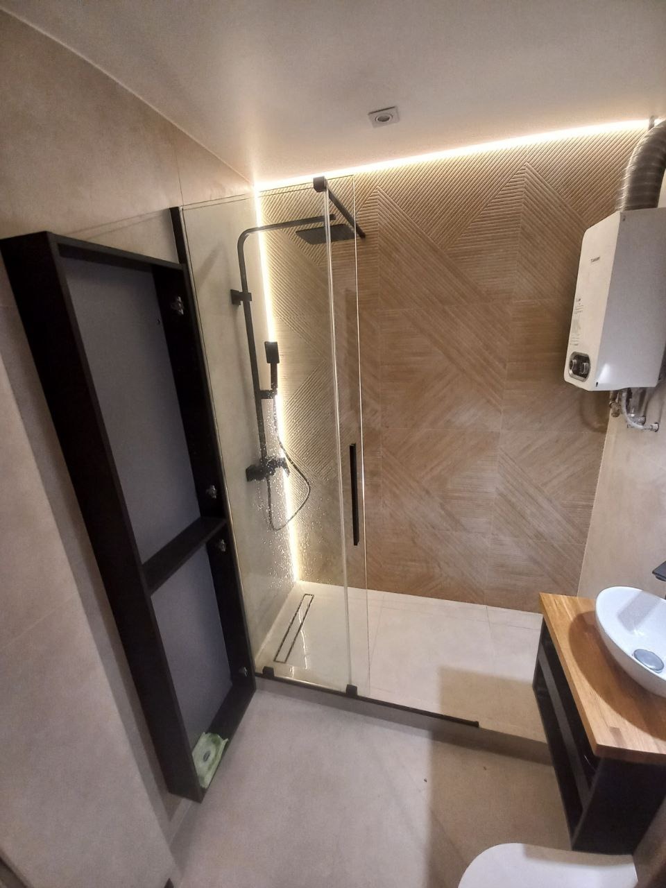 Nowoczesna łazienka z kabiną prysznicową typu walk-in z czarnymi profilami, podświetleniem LED za płytkami na ścianie oraz szafką z lustrem w kolorze ciemnego brązu i umywalką nablatową.