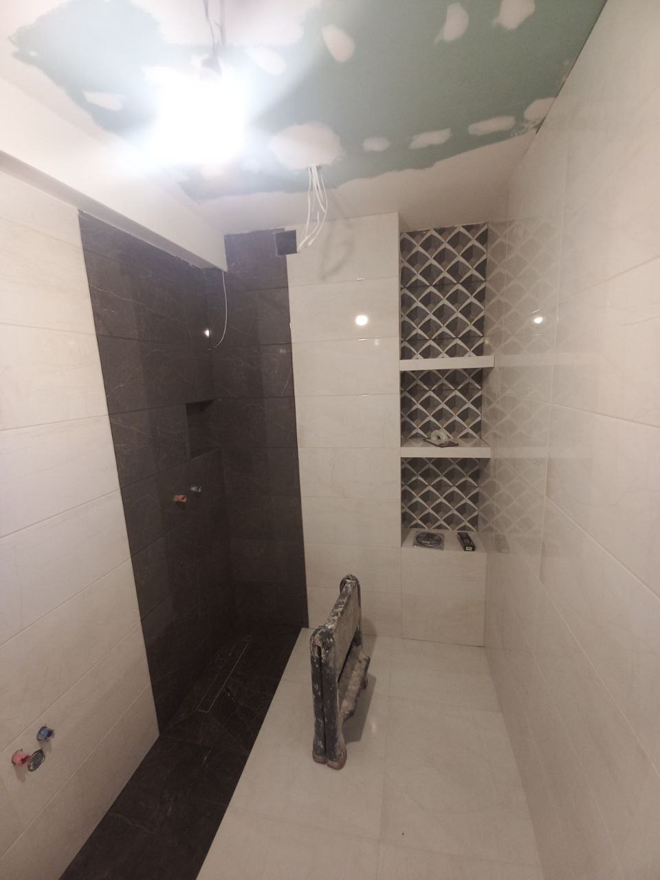 Wnętrze łazienki w trakcie remontu; widoczne płytki w odcieniach bieli i szarości, wnęka z dekoracyjnym wzorem, niezakończone prace na suficie i narzędzie glazurnicze.