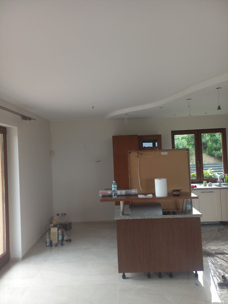 Wnętrze kuchni przed wykończeniem: świeżo pomalowane ściany i sufit z dekoracyjnym obniżeniem, widoczne butelki z wodą i materiały budowlane na podłodze, okno z widokiem na zieleń.