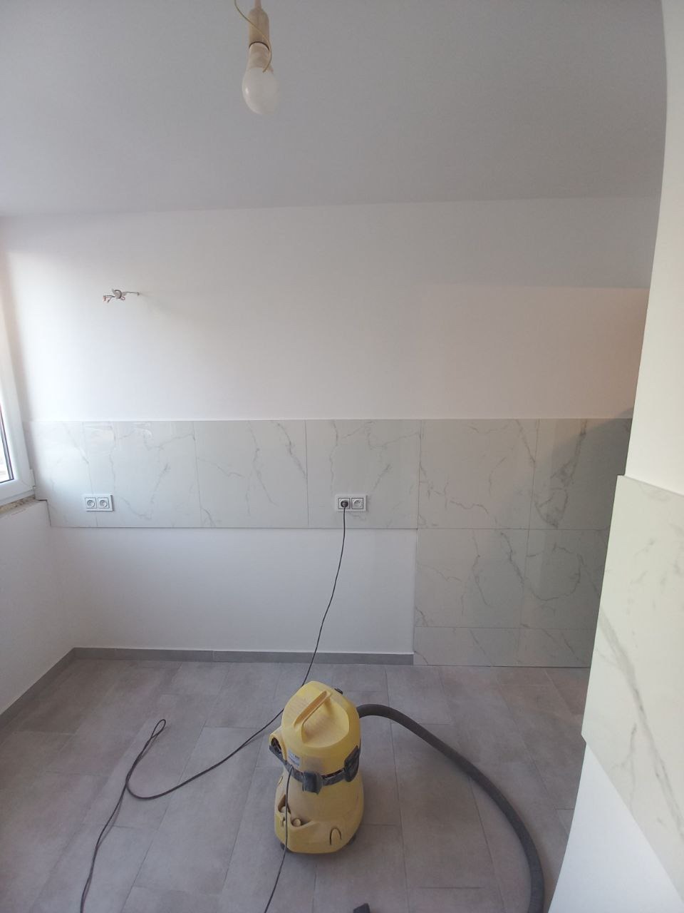 Wnętrze łazienki w trakcie remontu, z częściowo położonymi białymi płytkami imitującymi marmur na ścianie, szarą podłogą, żółtym odkurzaczem przemysłowym i gołą żarówką na suficie.