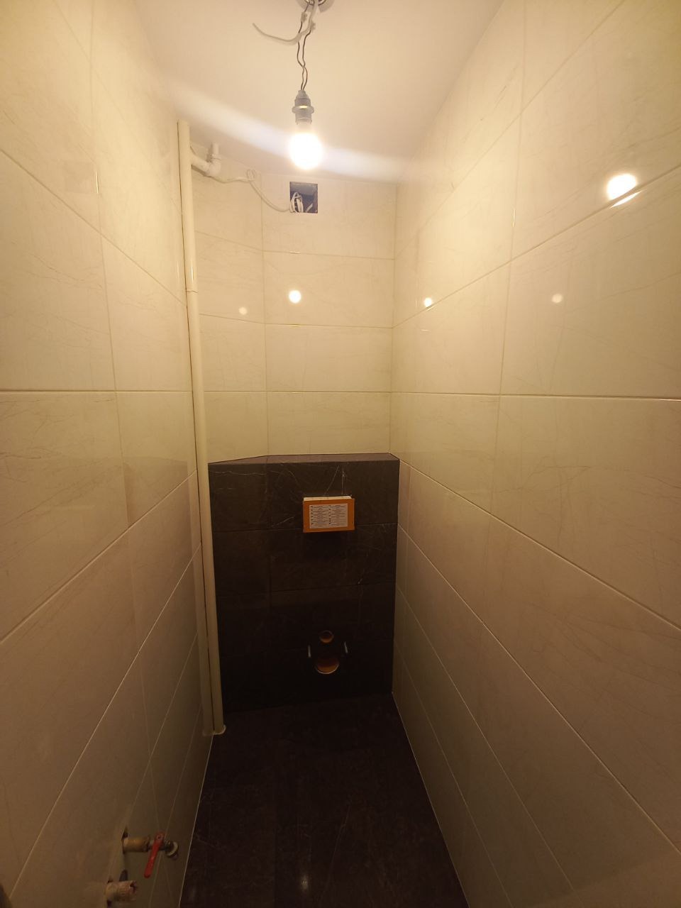 Wąska łazienka w trakcie remontu z beżowymi płytkami na ścianach, ciemną wnęką na toaletę i wystającą żarówką bez klosza.