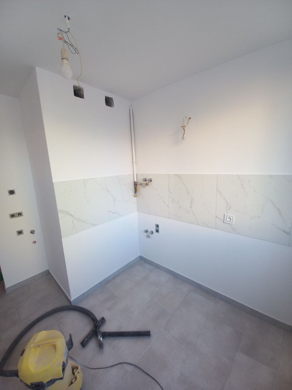 Wnętrze łazienki w trakcie wykańczania: częściowo położone płytki imitujące marmur, widoczne instalacje, surowa żarówka zwisająca z kabli, szary gres podłogowy oraz odkurzacz przemysłowy na podłodze.
