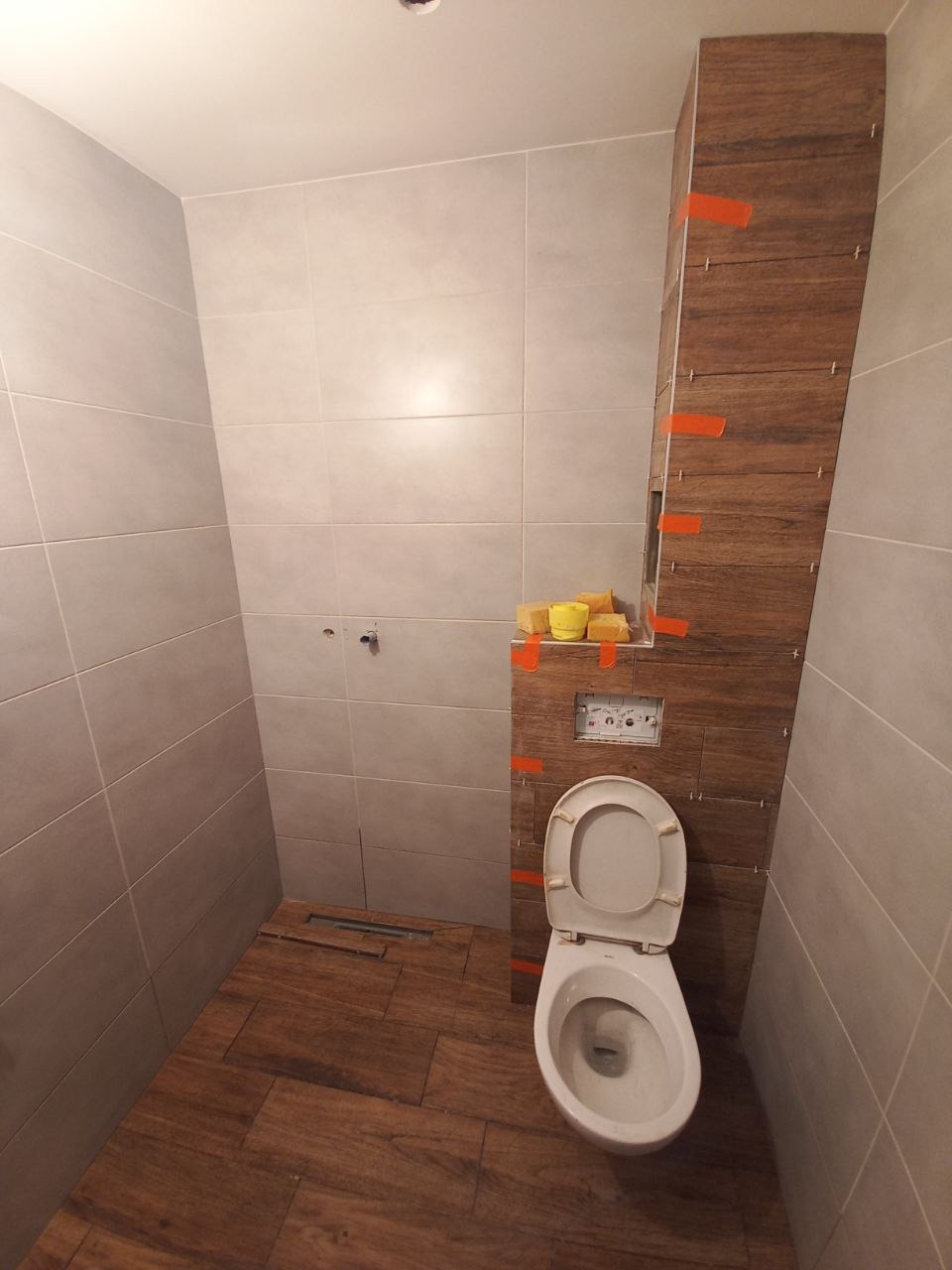 Wnętrze łazienki w trakcie wykańczania; widoczne płytki ceramiczne imitujące drewno na podłodze i częściowo na ścianie, białe płytki na pozostałych ścianach, zamontowana toaleta z otwartą klapą...