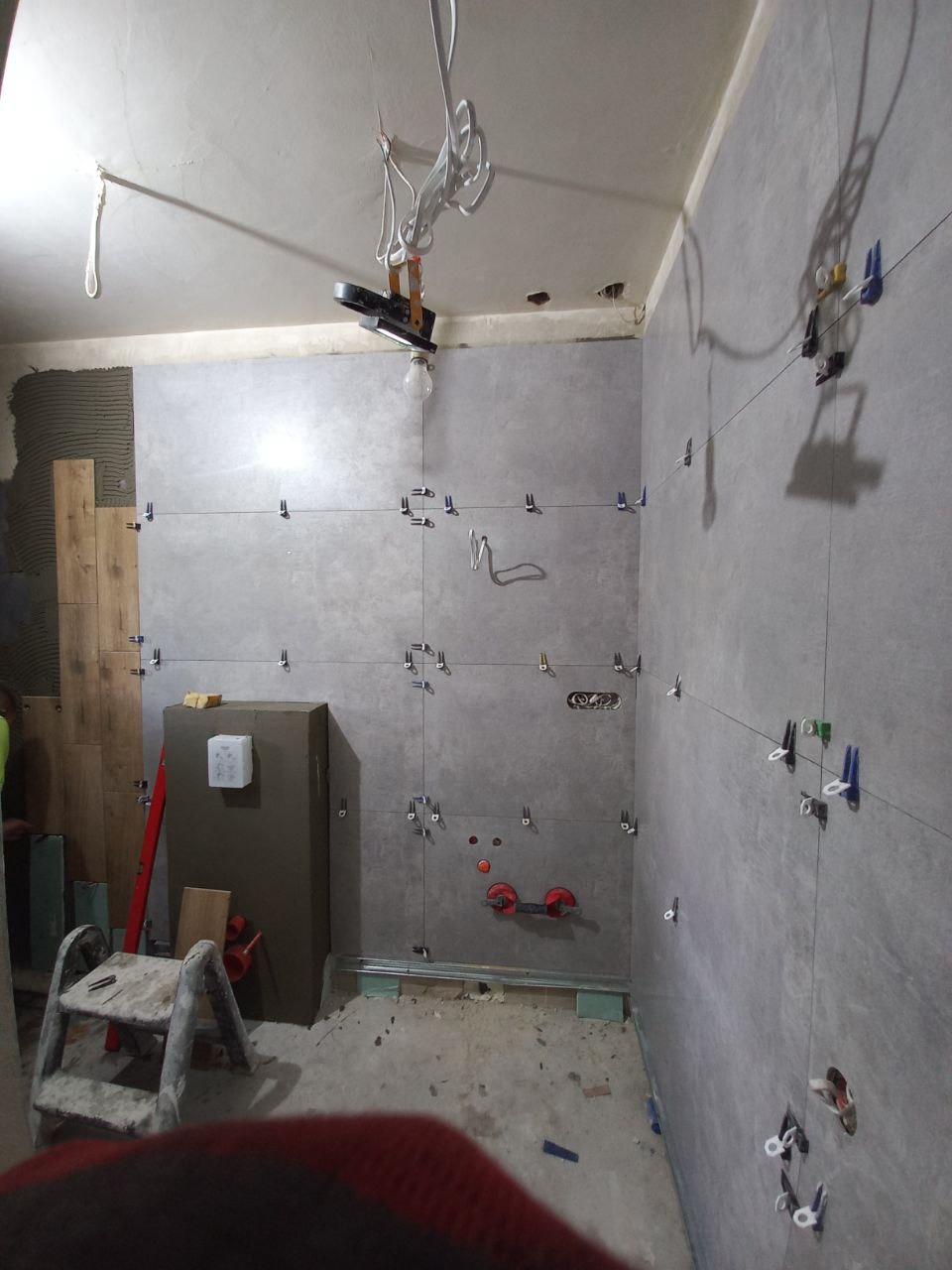 Układanie szarych płytek na ścianie w łazience w Katowicach, widoczne krzyżyki dystansowe, poziomica laserowa i drabina.