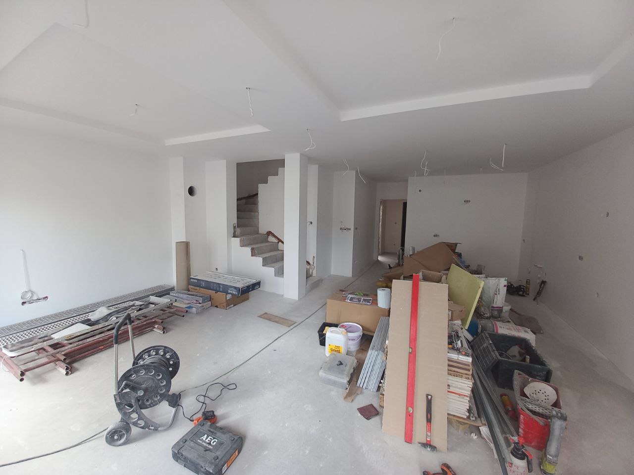 Wnętrze w trakcie remontu: pomalowane na biało ściany i sufit, widoczne schody w surowym stanie, narzędzia i materiały budowlane rozłożone na podłodze, wystające kable ze ściany i sufitu.