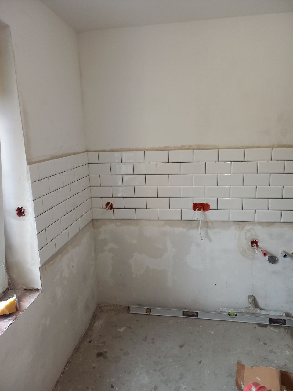 Fragment ściany z ułożonymi białymi, prostokątnymi płytkami, częściowo wykończona powierzchnia, widoczne puszki elektryczne, poziomica na podłodze, etap prac wykończeniowych.