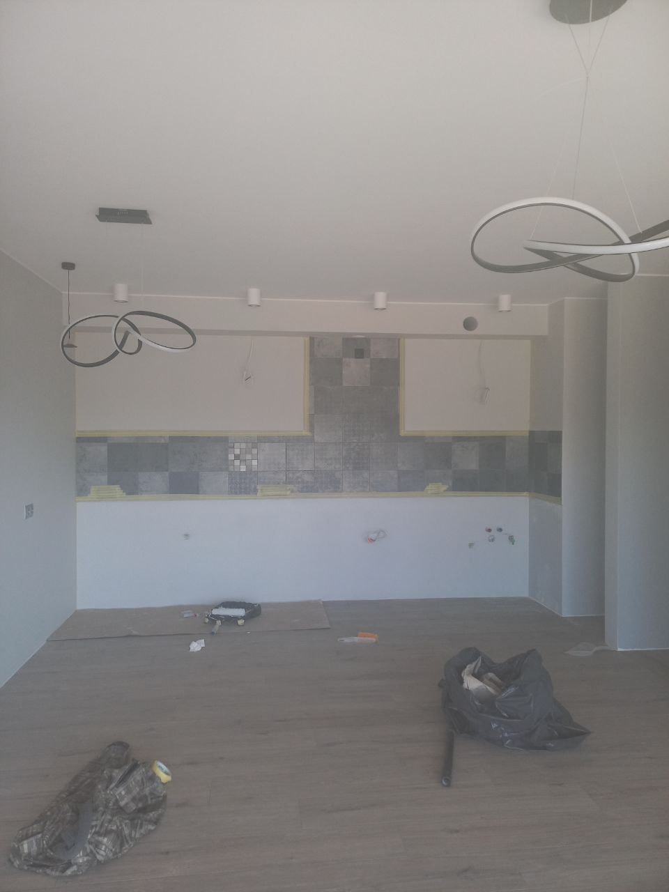 Wnętrze w trakcie remontu: świeżo położone szare płytki na ścianie zabezpieczone taśmą malarską, widoczne przewody, na podłodze narzędzia i worek na śmieci, nowoczesne lampy sufitowe.