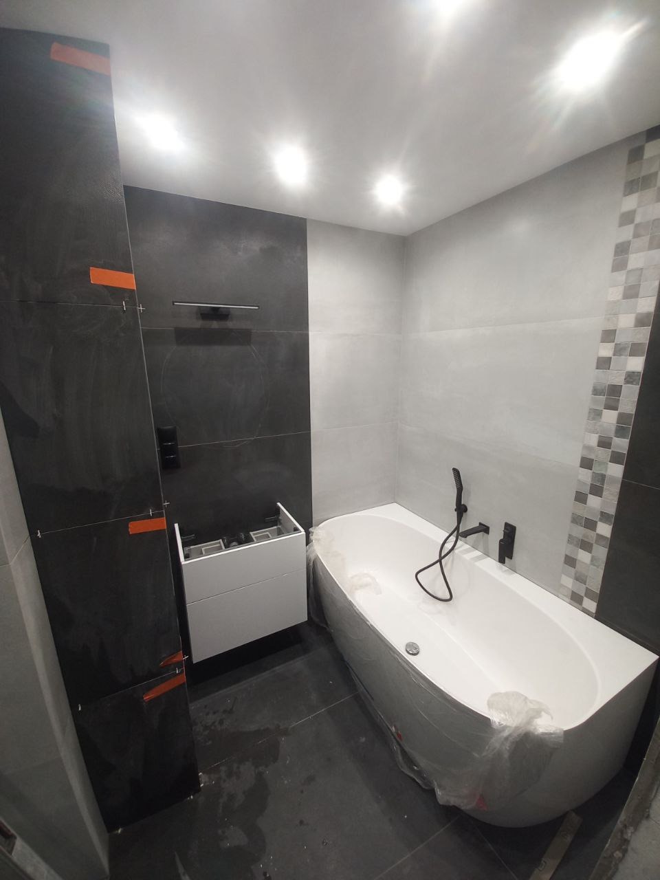 Nowoczesna łazienka w trakcie wykańczania: czarne płytki na ścianie z pomarańczowymi taśmami, biała wanna owinięta folią ochronną, biała szafka pod umywalkę bez umywalki, szare płytki na podłodze...